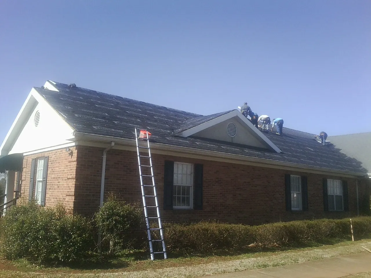 Crew tearing shingles off a brick ranch roof with a ladder set — Emergency Roof Repair work by our crew in Alvord