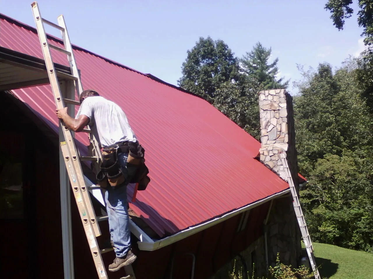 Roofer on a ladder beside a red metal roof and stone chimney — Soffit & Fascia Repair by Roofing Brothers in Alvord